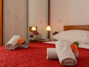 Une chambre d’hôtel avec une couvre-lit à motifs rouges, deux serviettes roulées ornées de roses orange, une tête de lit en bois, un oreiller blanc et deux lampes de chevet.