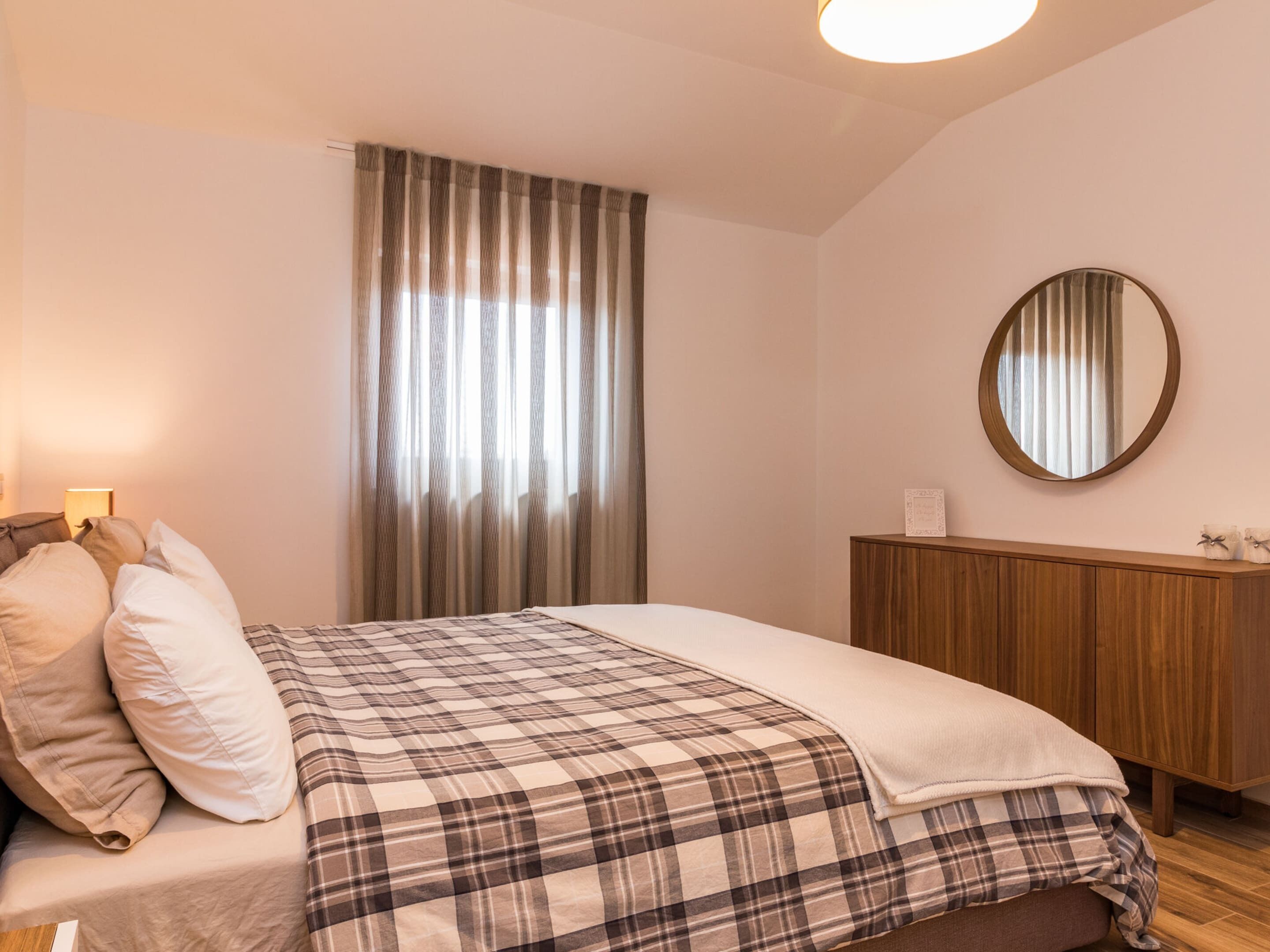 A bedroom with a bed, a wooden dresser, a round mirror, and a window with curtains.