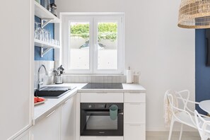 Cocina compacta con horno integrado, ventana con vistas a zonas verdes y una silla blanca.