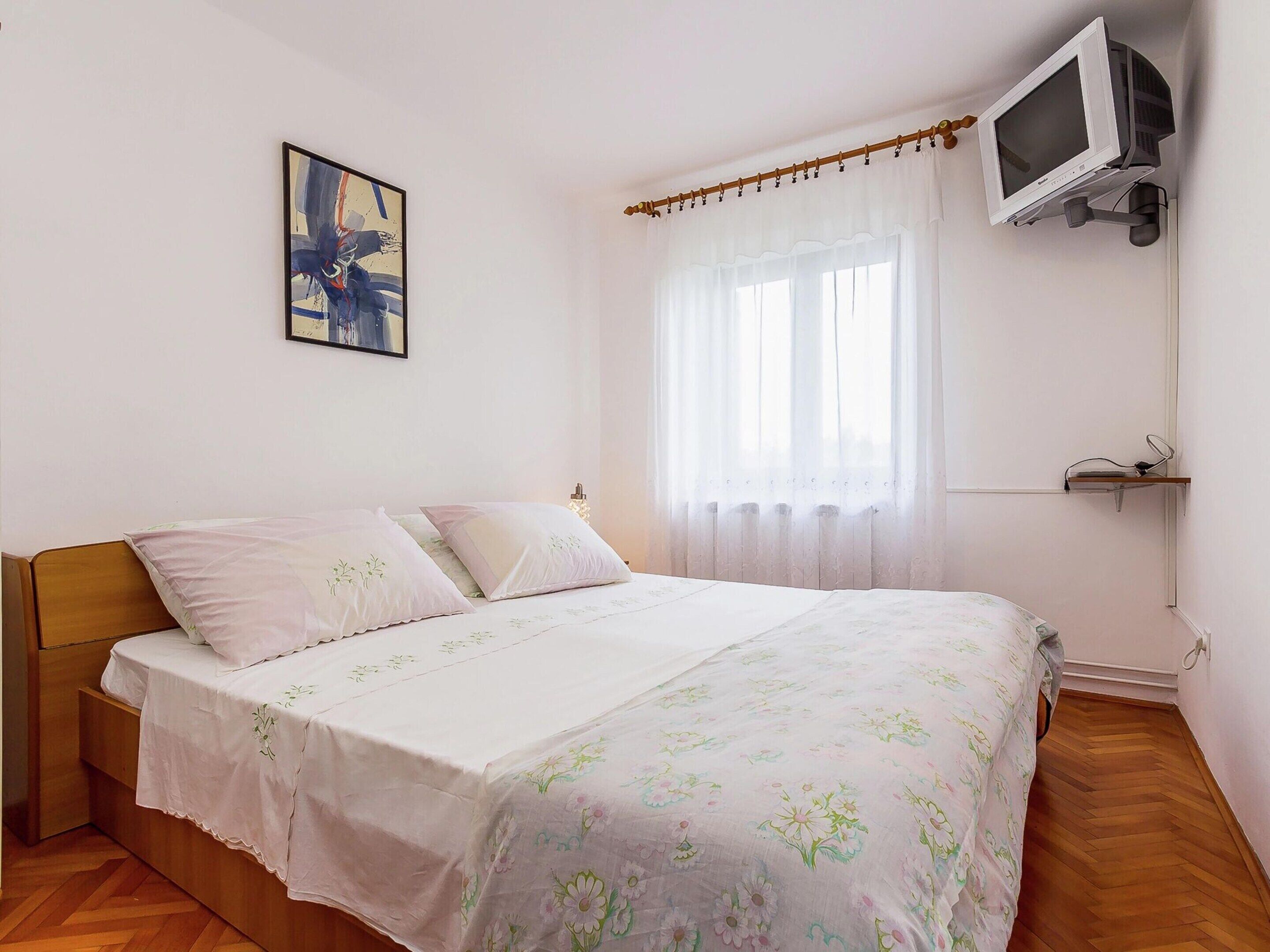 A bedroom with a bed, a TV mounted on the wall, a window with curtains, and a painting on the wall.