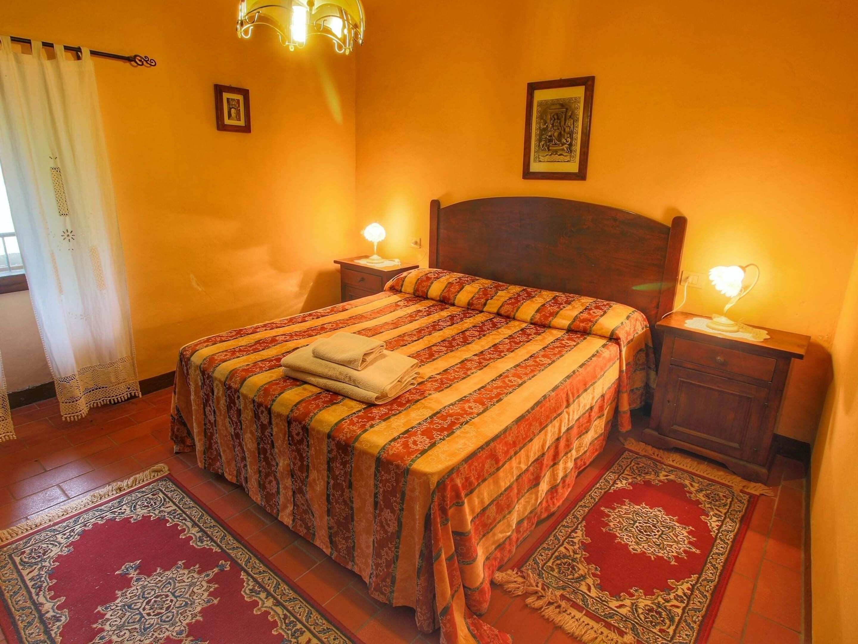 A bedroom with a large bed, two bedside tables, a nightstand, a patterned rug, and a window with curtains.