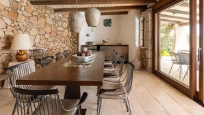 A dining area with a stone wall, wooden ceiling beams, hanging light fixtures, and a view through large windows. - Luxe Cote Stay- Elegant Villa (Arzachena)