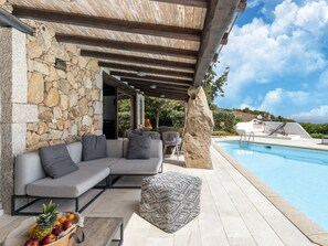 An outdoor pool area with a stone wall, a gray sofa with cushions, a patterned ottoman, and a fruit basket. - Luxe Cote Stay- Elegant Villa (Arzachena)