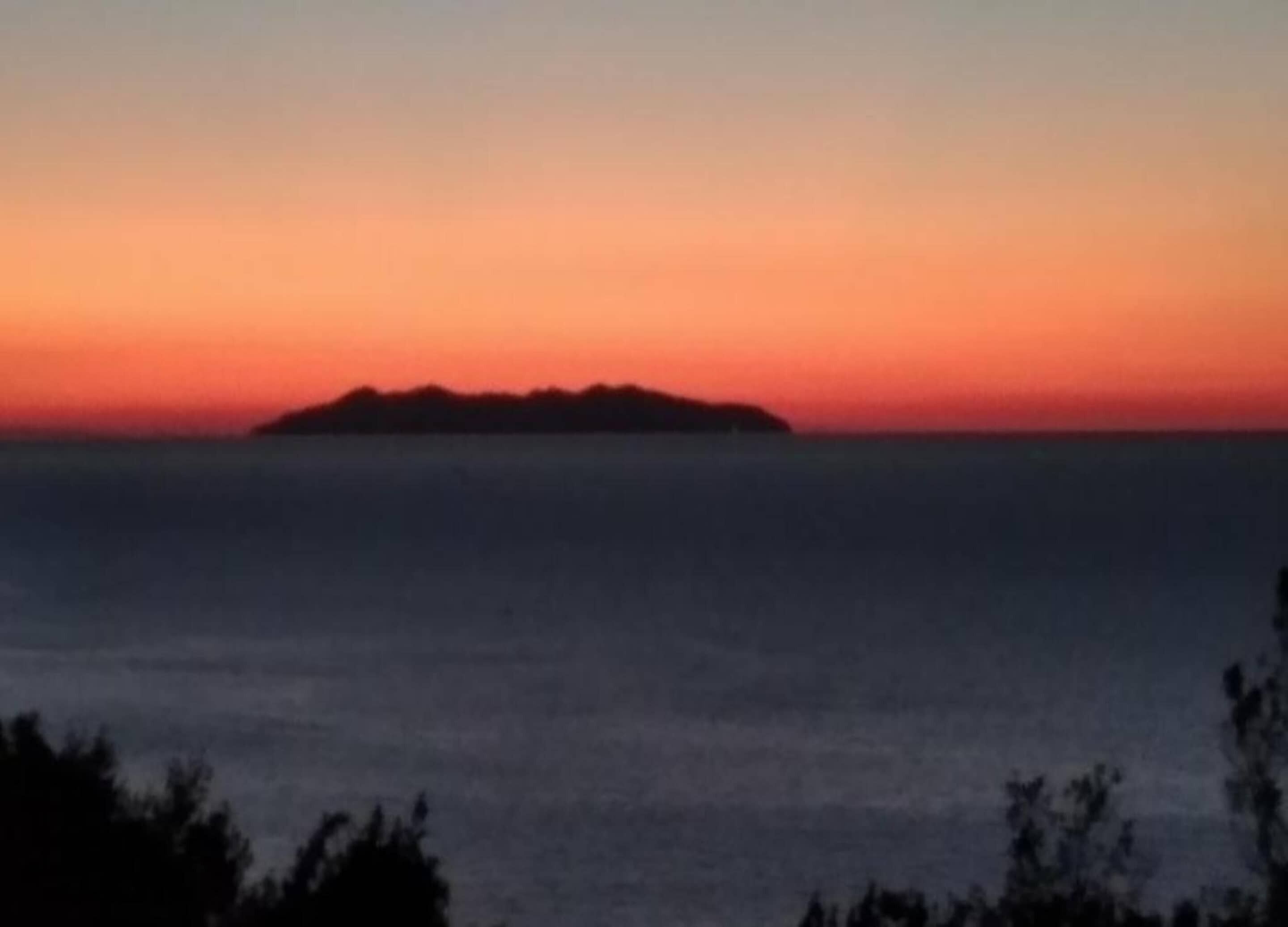 Auringonlaskun aikaan avautuva näkymä pienelle saarelle, jonka ympärillä vesi on tyyntä.