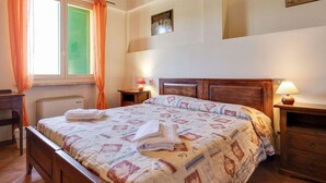 A bedroom with a wooden bed, a nightstand with a lamp, a window with curtains, and a painting on the wall. - Mansion Stay With Scenic Views (Siena)