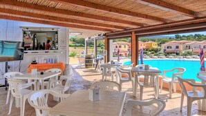 A covered outdoor seating area with white plastic chairs and tables, a bar counter, and a view of a swimming pool. - Belvilla by OYO Sardegna Mare 617 (Trinità d'Agultu e Vignola)