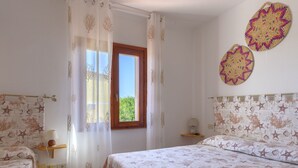 A bedroom with two beds, a window with curtains, a bedside table with a lamp, and wall decorations. - Belvilla by OYO Sardegna Mare 617 (Trinità d'Agultu e Vignola)