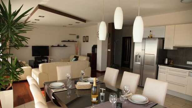 Un espace repas moderne avec une table en bois foncé, dressée pour quatre personnes, entourée de chaises blanches. Une grande plante en pot est visible sur la gauche, et la pièce présente une palette de couleurs neutres, avec des murs blancs et un plafond foncé.