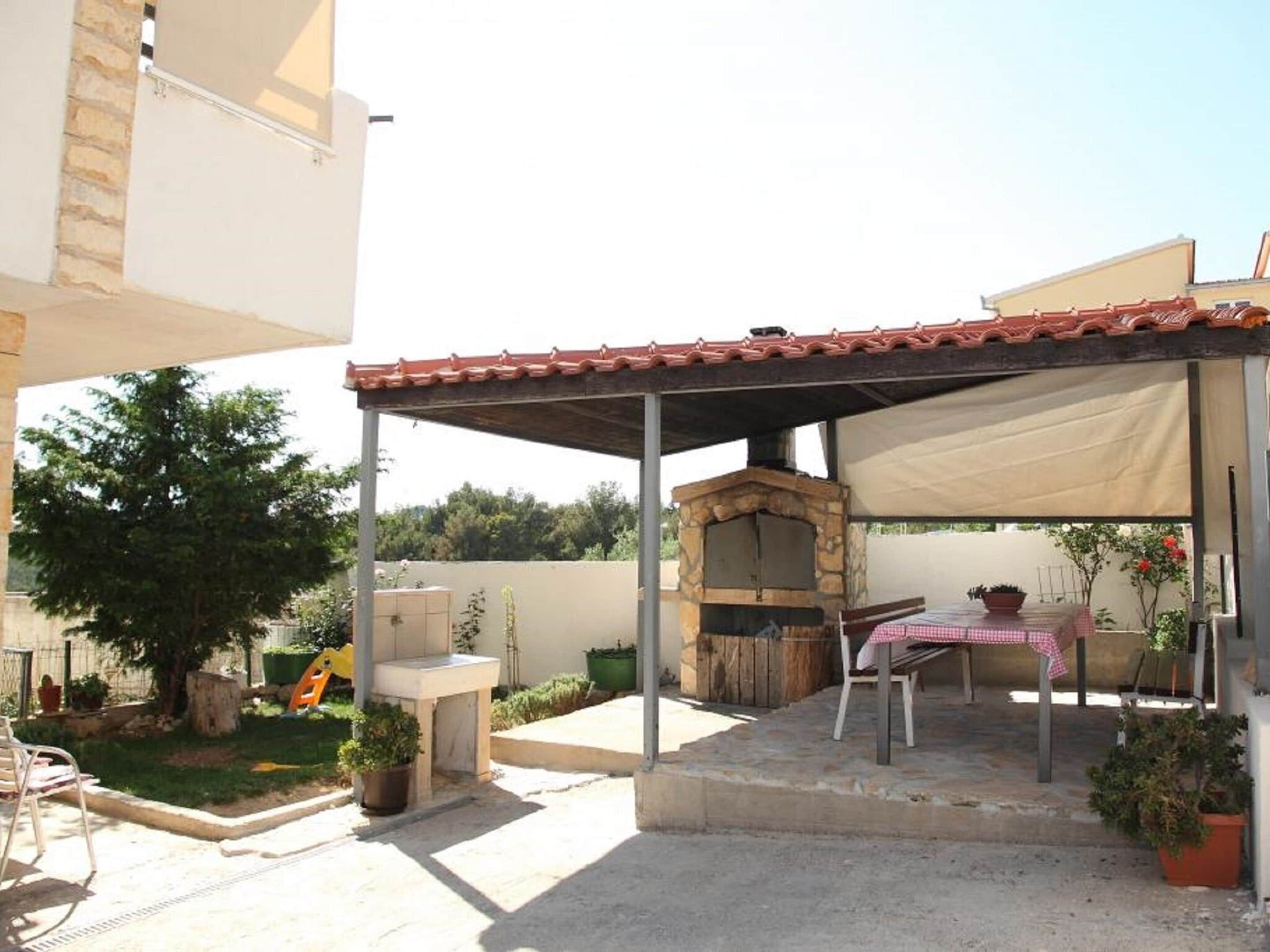 A covered outdoor area with a stone-built oven, a table with a checkered tablecloth, and potted plants.