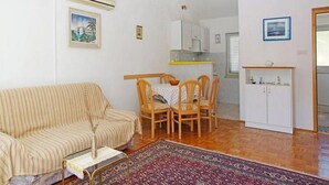 A living room with a striped sofa, a dining table with chairs, a patterned rug, and a kitchen area with cabinets and a window. - Apartments Tomasic - Two Bedroom Apartment With Terrace and Sea View (Smokvica)
