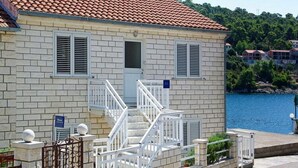 A two-story building with a tiled roof, a staircase with white railings, and a view of a body of water. - Apartments Tomasic - Two Bedroom Apartment With Terrace and Sea View (Smokvica)