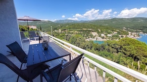 A balcony with a table and chairs, overlooking a coastal landscape with mountains and a town.