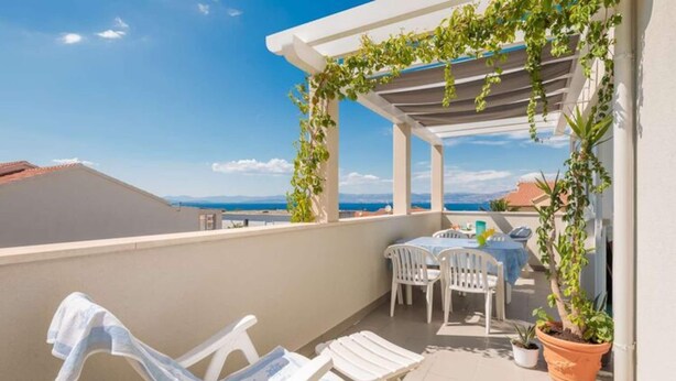 Een balkon met witte stoelen en een tafel, planten in potten, en uitzicht op zee.