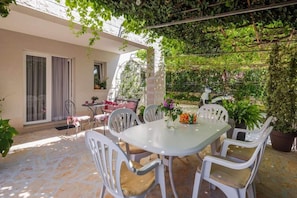 En udendørs terrasse med et bord og stole, omgivet af krukker med planter og en espalierbeklædt væg.