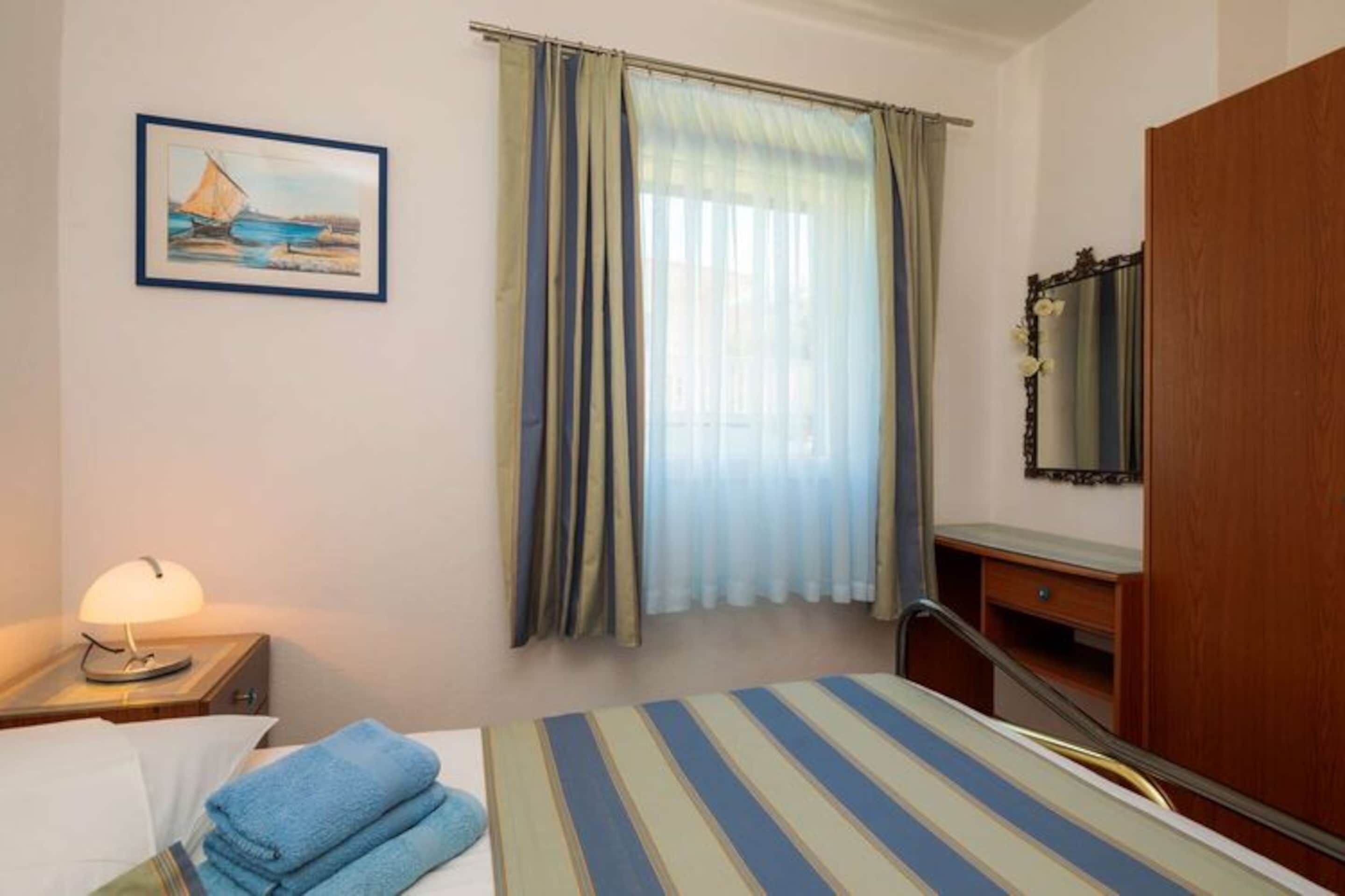 Une chambre d’hôtel comprenant un lit, une table de chevet avec une lampe, une commode en bois, un miroir et une fenêtre avec des rideaux.