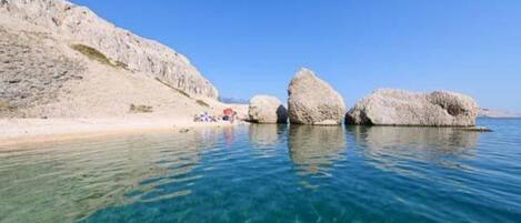 A clear coastal scene with three large rock formations in the water and a sandy beach with a few umbrellas.