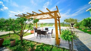 En træpergola med et bord og stole, en swimmingpool i baggrunden og en velholdt have med plantekasser.
