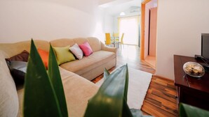 Ein Wohnzimmer mit einem beigefarbenen Sofa, einem Fernseher auf einem Holzboden und einem Essbereich im Hintergrund.