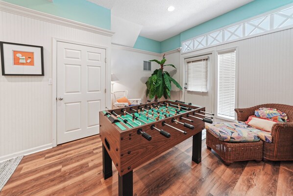 A room with a foosball table, a wicker sofa, and a door with a plant beside it.