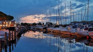 Ein Yachthafen mit Booten, die an Holzstegen liegen, ruhiges Wasser, das Boote und Himmel reflektiert, und Gebäude mit Lichtern im Hintergrund.