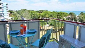 Ein Balkon mit Tisch für zwei Personen, einem Ventilator und Blick auf Meer und Grünfläche.