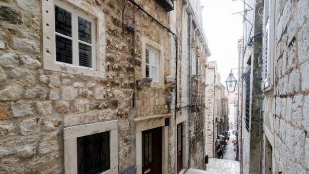 Une ruelle étroite pavée, bordée de bâtiments des deux côtés, un petit escalier menant à une porte, et un lampadaire au loin.