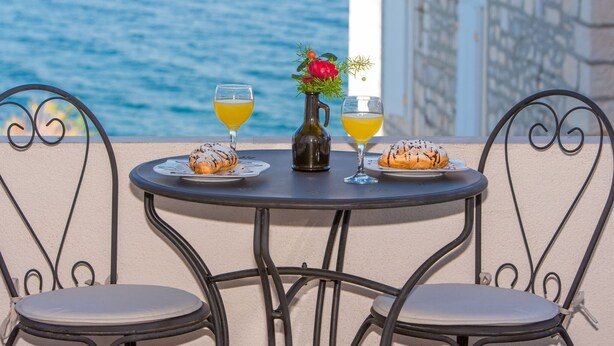 A table with two chairs, a bottle, and two glasses of orange juice, set against a backdrop of a body of water and a building. - Apartments Tino- Two Bedroom Apartment With Balcony and Sea View (Sutivan)