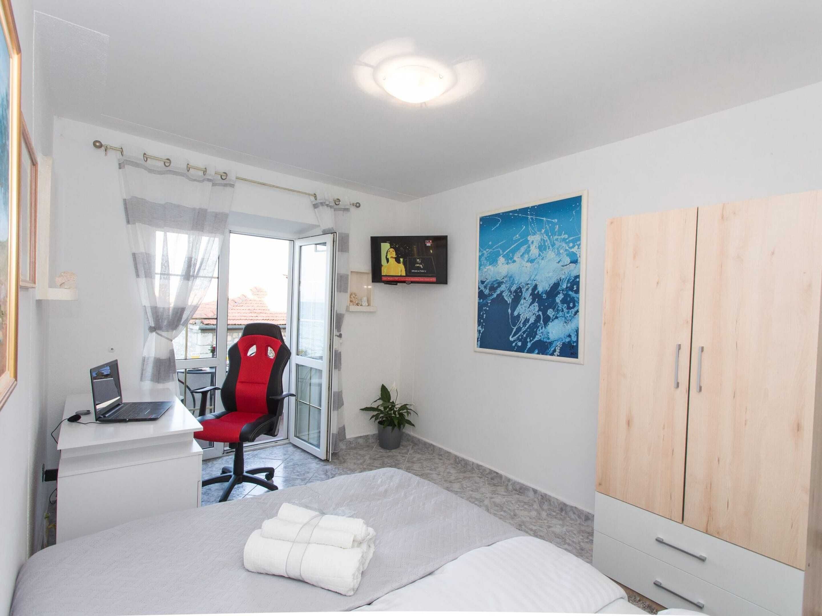 A bedroom with a bed, a desk with a laptop, a red chair, a wooden wardrobe, a potted plant, and a large abstract painting on the wall.