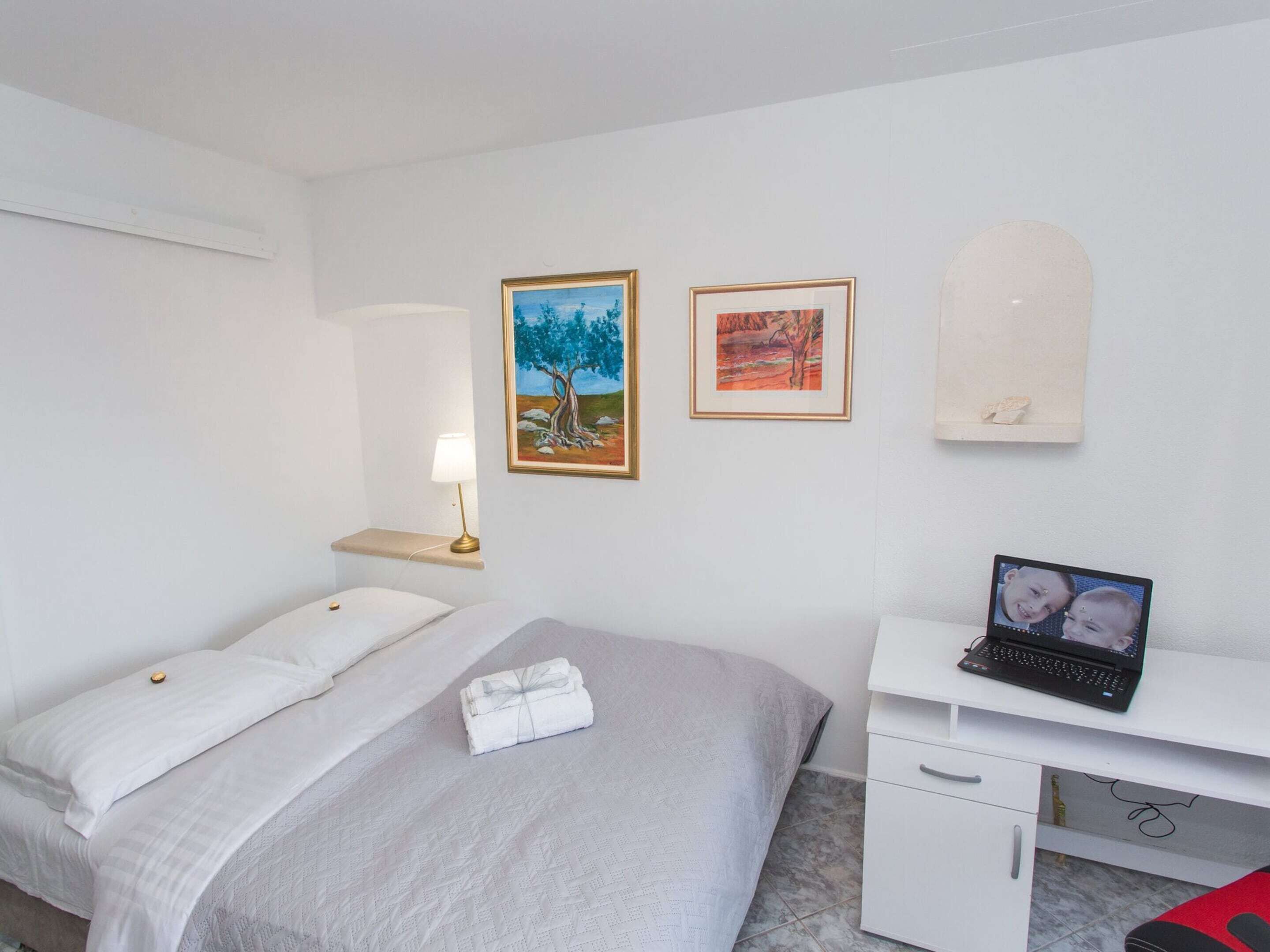 A hotel room with a bed, a desk with a laptop, and two framed pictures on the wall.