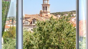 A view of a building with a bell tower and red-tiled roofs through a window with a green bush in the foreground. - Apartment Marcela - Deluxe Two Bedroom Apartment With Terrace and Sea View (Sutivan)