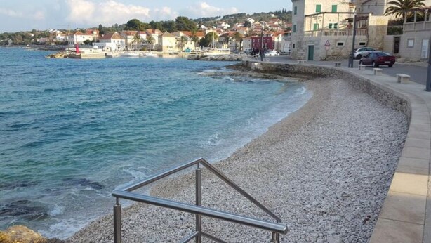 A coastal scene with a pebble beach, a metal railing, and buildings in the background. - Apartment Marcela - Deluxe Two Bedroom Apartment With Terrace and Sea View (Sutivan)