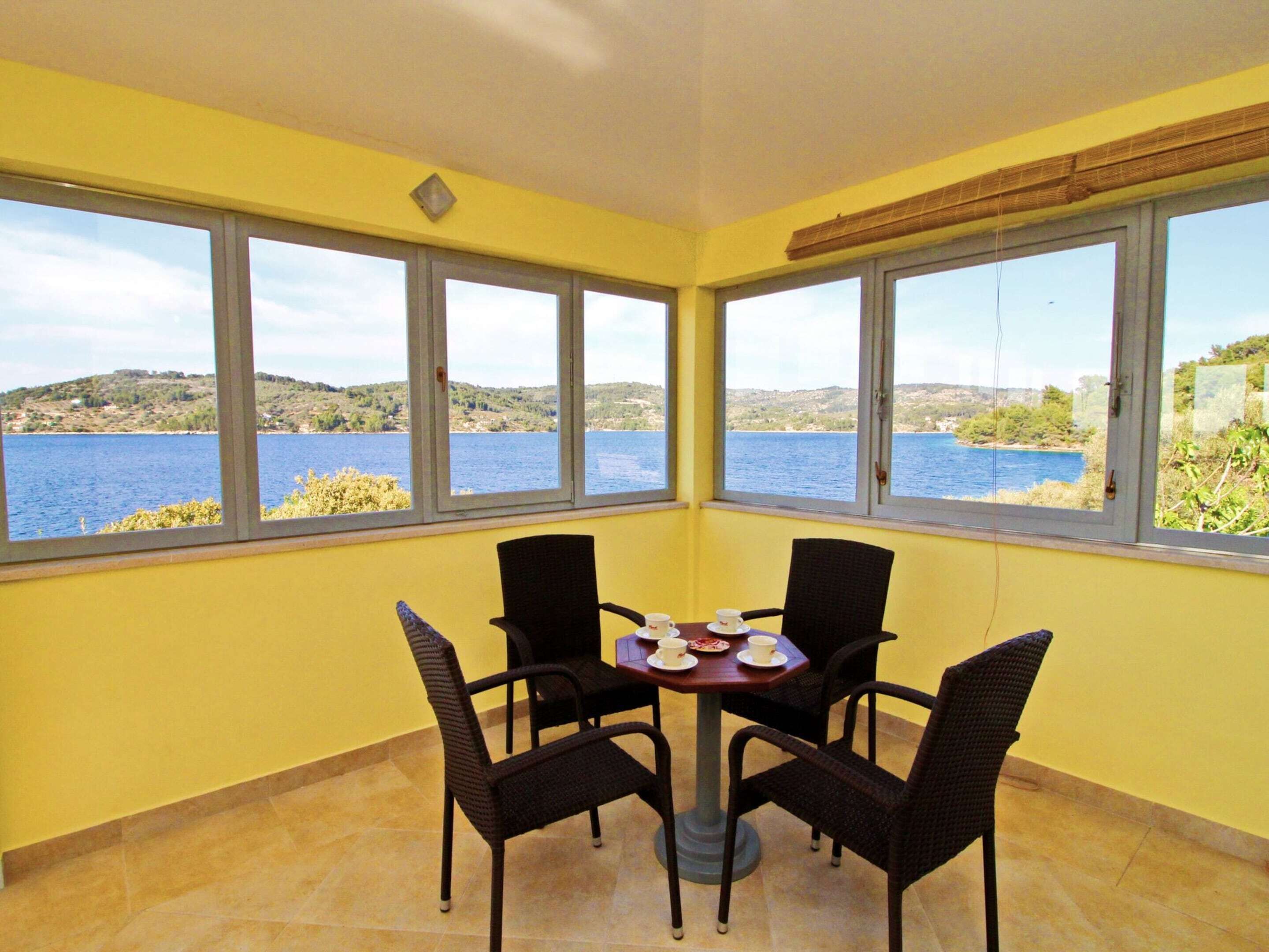Ein Zimmer mit Tisch und vier Stühlen, großen Fenstern mit Blick auf einen See und Hügel, gelben Wänden und Fliesenboden.