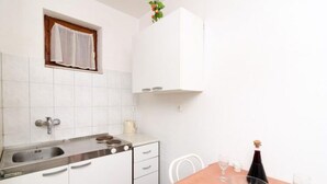 A small kitchen with a sink, stove, and cabinets. There is a window with a curtain, a plant, and a table with a bottle and glasses.