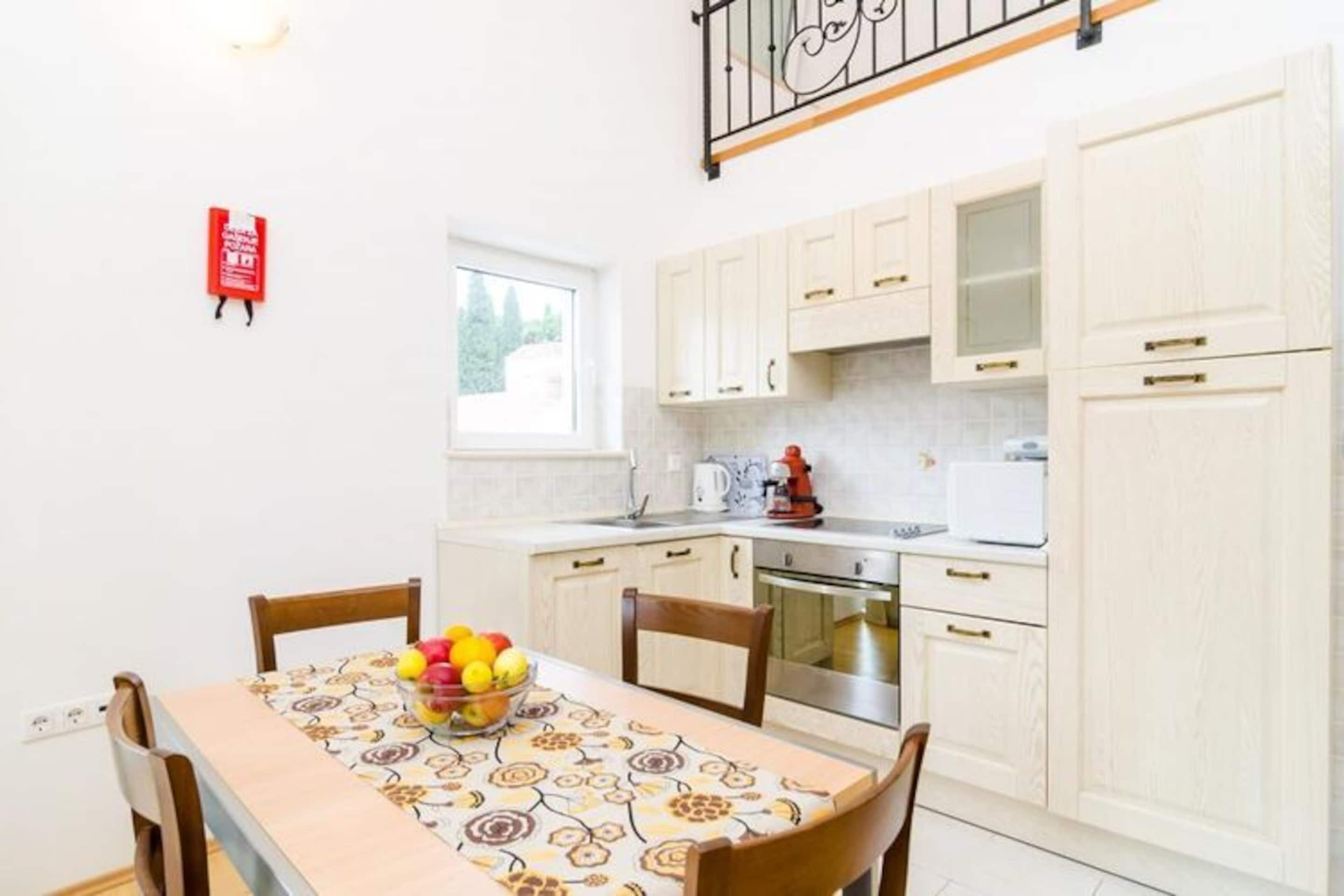 A compact kitchen with white cabinets, a stainless steel oven, and a microwave. A fruit bowl is on the table, and a fire extinguisher is mounted on the wall.