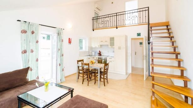 A bright living room with a sofa, a glass coffee table, and a dining area with a table and chairs. There is a staircase with a railing leading to an upper floor. - Apartments Nikola - Comfort One Bedroom Apartment With City View (Dubrovnik)