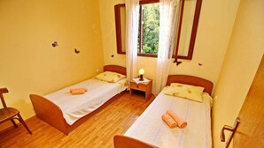 A small, clean room with two single beds, wooden flooring, a window with curtains, and a nightstand with a lamp.