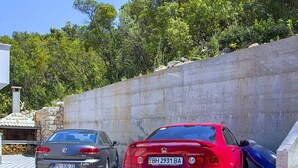 To biler parkeret i et overdækket parkeringsområde med en stenvæg og træer i baggrunden.