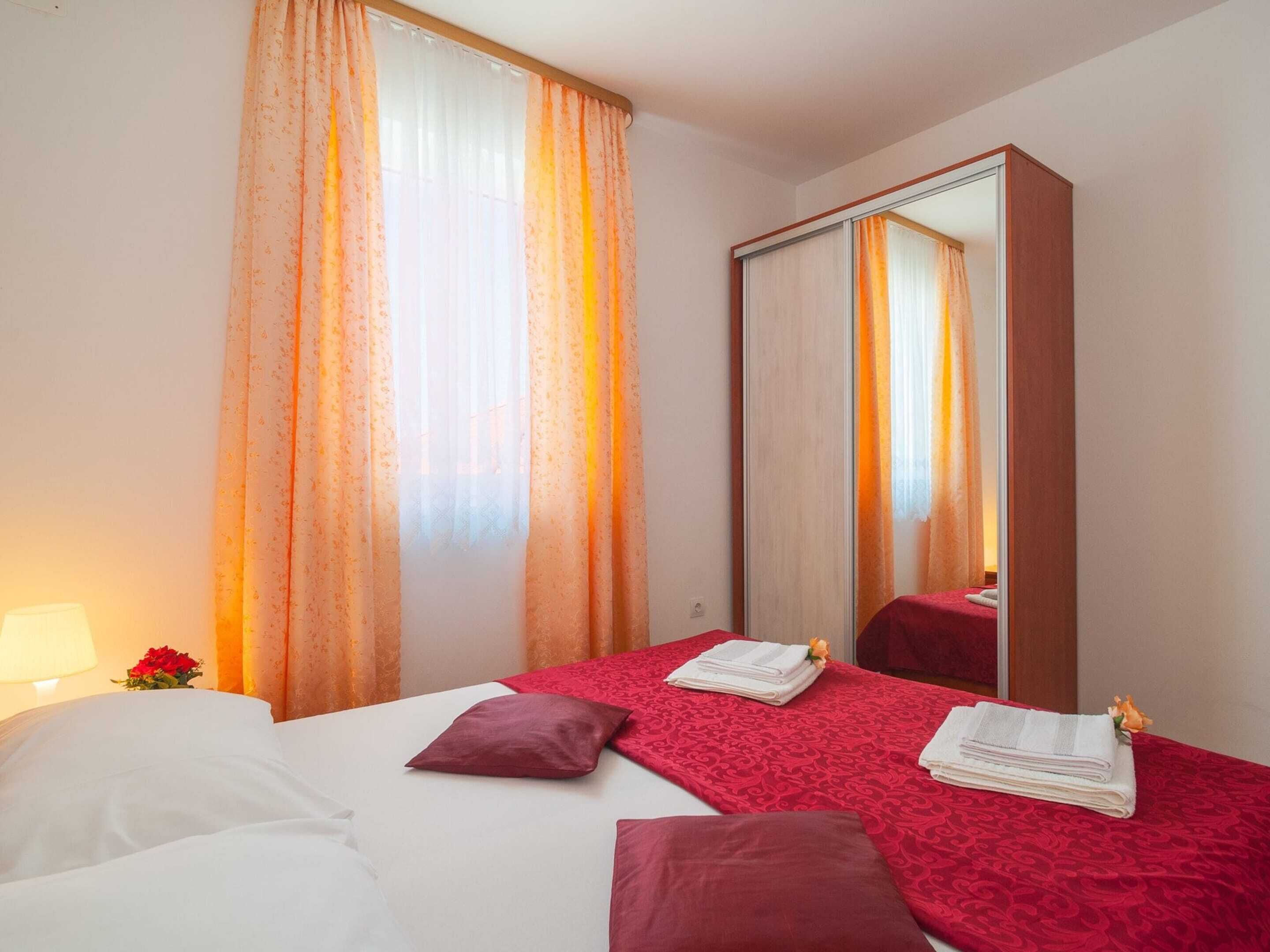 A hotel room with a bed, red and white bedding, a large mirror, and a window with orange curtains.