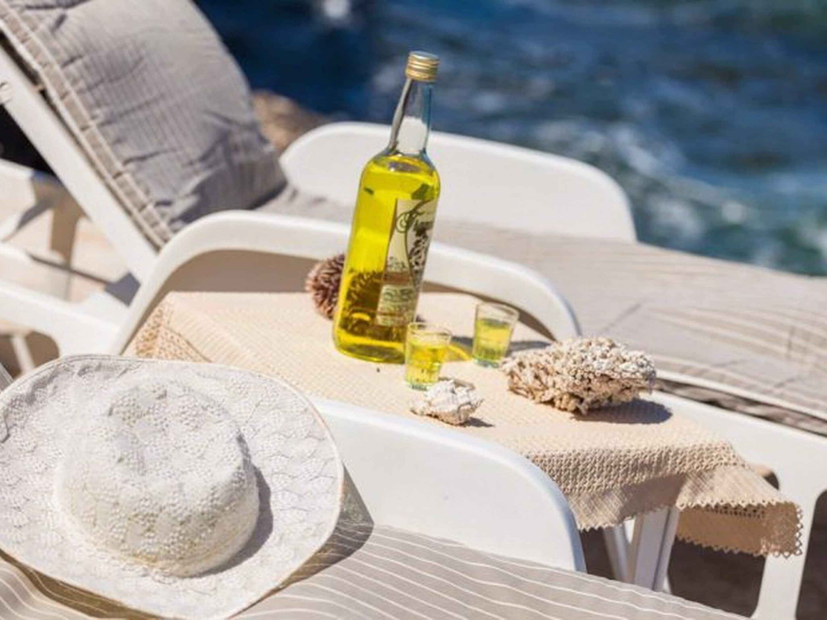 A sun lounger with a bottle of olive oil, a glass, and a hat on a table by a pool.