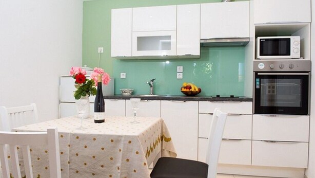 A compact kitchen with white cabinets, a microwave, and an oven. A dining table with a floral tablecloth and chairs is set up in the center. - Apartments Stella - One Bedroom Apartment With Terrace A2 (Dubrovnik)