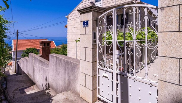 A gated entrance with a metal gate, a concrete wall, and a building with a tiled roof. - Apartments Stella - One Bedroom Apartment With Terrace A2 (Dubrovnik)