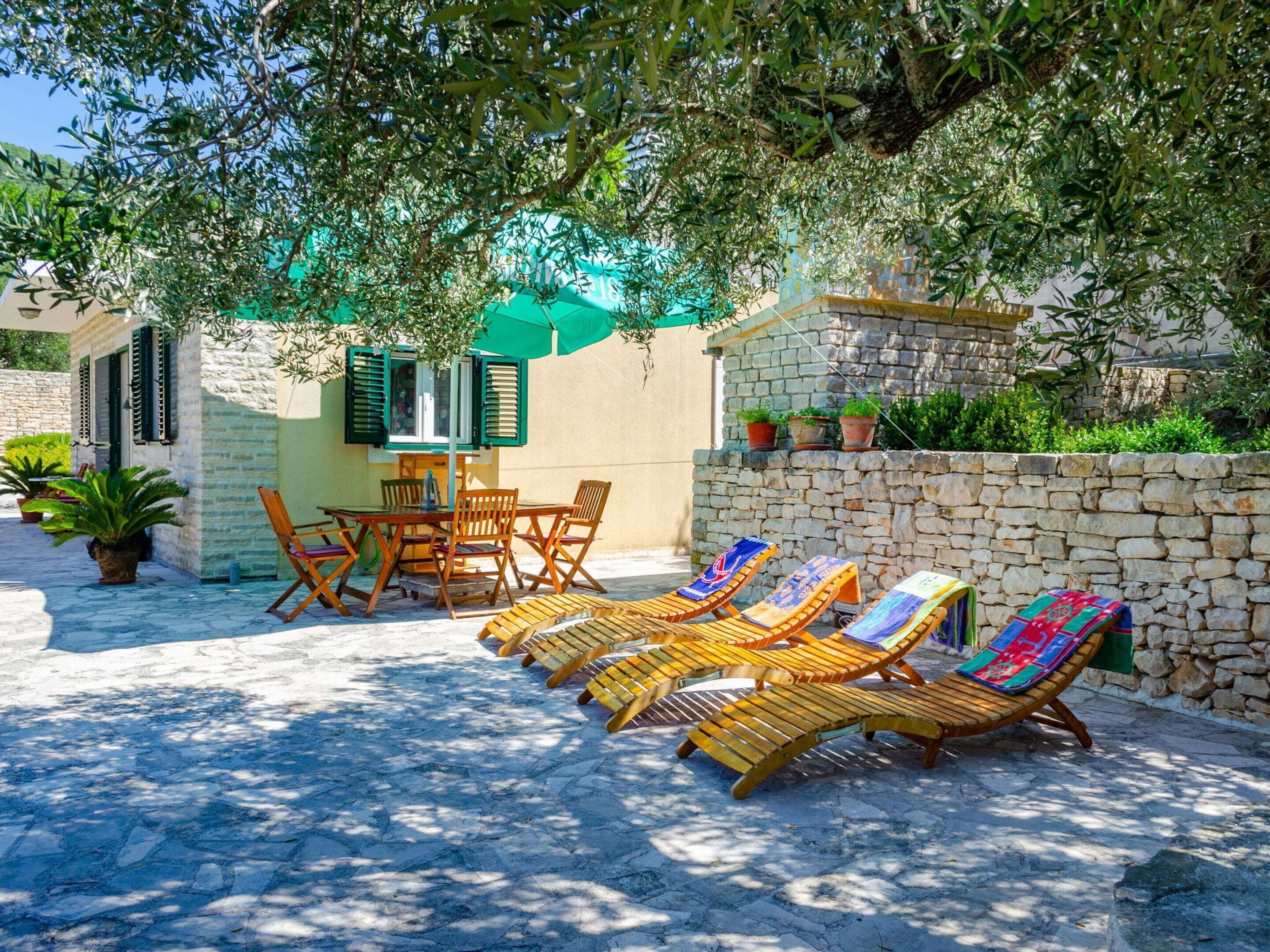 A sunlit outdoor seating area with wooden lounge chairs and a table, surrounded by stone walls and greenery.