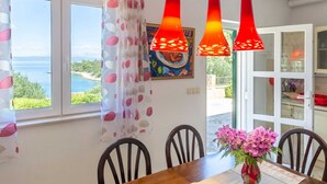 A dining area with a wooden table, chairs, a vase of flowers, and a view of the sea through large windows. - Vacation House Marija, Korcula-two Bedroom Holiday Home With Terrace and Sea View (Korcula)