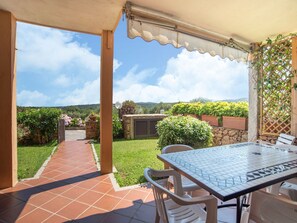 A covered patio with a tiled floor, a table with chairs, and a view of a garden and hills in the background. - Belvilla by OYO Monolocale Baia 8 (Golfo Aranci)