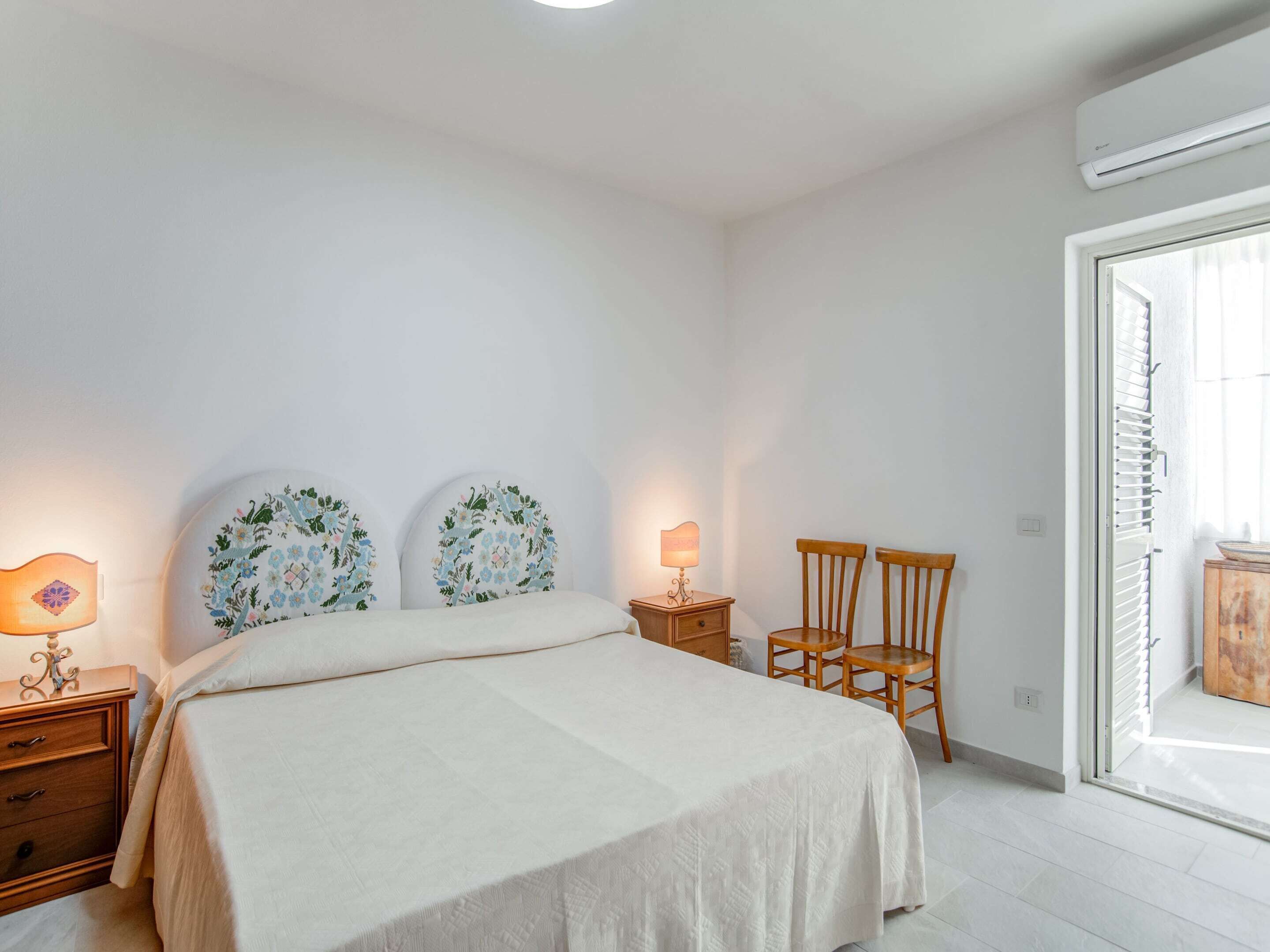 Un dormitorio con una cama, dos mesitas de noche, una lámpara, dos sillas y un ventanal con vistas.
