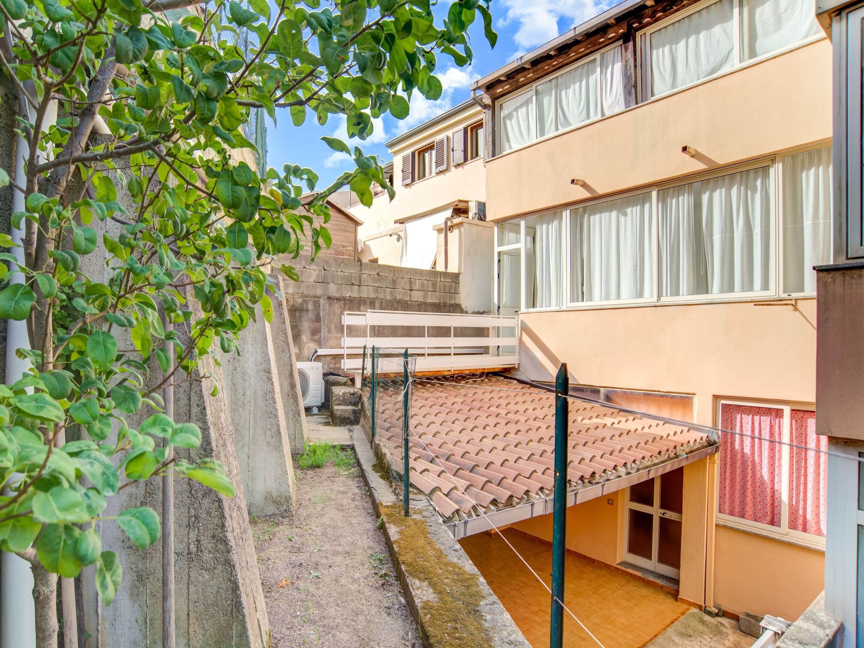 A residential building with a tiled patio, a concrete pathway, and a small garden area with a tree.