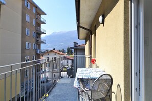 Un balcon étroit avec une table et des chaises, une balustrade et une vue sur des bâtiments et des montagnes en arrière-plan.