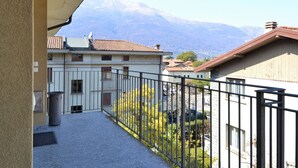 A balcony with a view of a mountain range, a building with a tiled roof, and a fenced area with greenery. - Apartment by Lake Como With Scenic Views (Dervio)