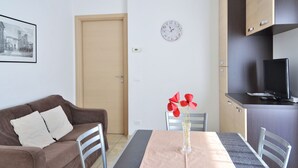 A small, modern apartment with a brown sofa, a dining table with a vase of red flowers, a clock, and a TV on a cabinet. - Apartment by Lake Como With Scenic Views (Dervio)