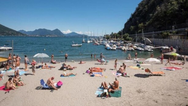 A beach with people sunbathing and swimming, boats docked in the background, and a mountainous landscape. - Pleasant Apartment by the Lake (Baveno)